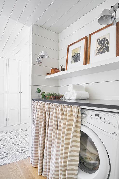 Short beige stripe curtains hid a white front load washer and dryer with a vintage style. Shiplap walls invite a cottage appeal along with a white floating shelf above the washer and dryer set.