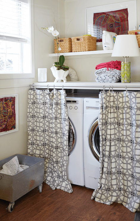 Lake house laundry room features a white front load washer and dryer tucked under a countertop hidden behind curtains. A washer and dryer stands under a floating shelf filled with woven baskets alongside a galvanized metal rolling laundry cart.