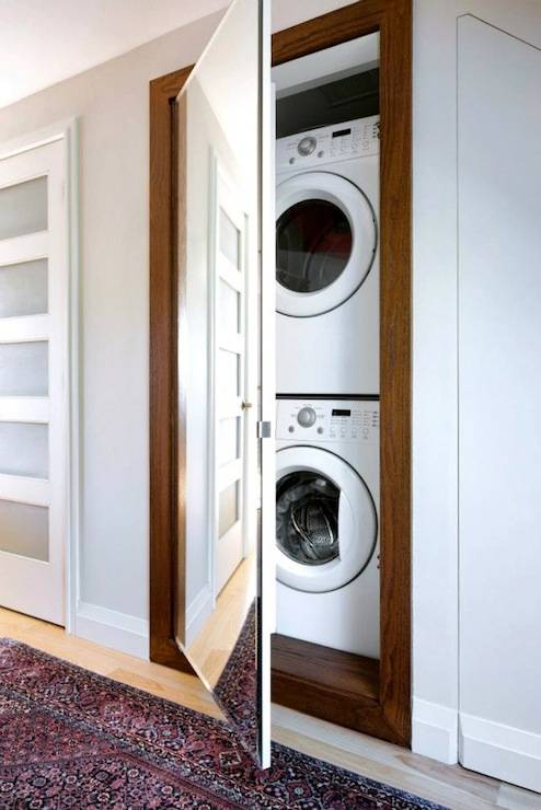Laundry area with stacked washer and dryer hidden behind a custom framed out mirror on a touch latch that when closed looks simply like a floor length mirror. The hidden laundry area in the hallway is framed by gray walls over light hardwood floors topped with a Persian-style rug.