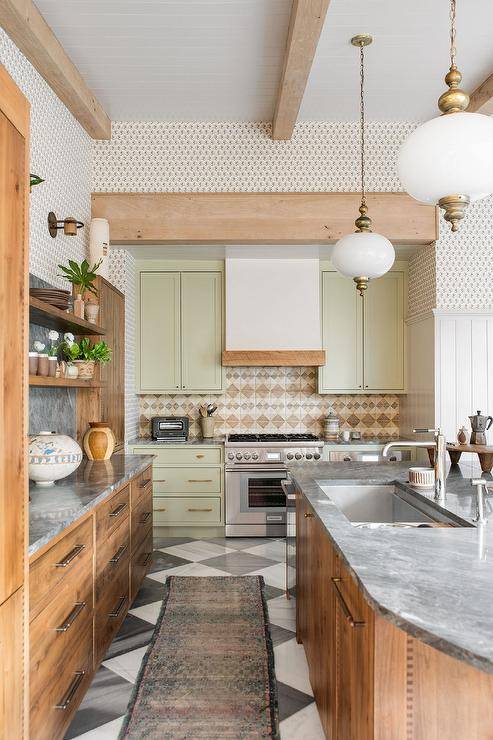 Cottage kitchen features pale green cabinets with tan backsplash tiles, reclaimed oak cabinetry topped with gray marble, a rustic oval island with dark gray marble countertop and island sink illuminated by glass lanterns and black and white Harlequin floor tiles.
