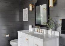 White glass and brass sconces are mounted flanking a black French vanity mirror hung over a white bath vanity fitted with a slatted shelf, brass hardware, and an aged brass faucet kit. The washstand sits on taupe and black geometric floor tiles beside a white porcelain toilet fixed under a black and white art piece.