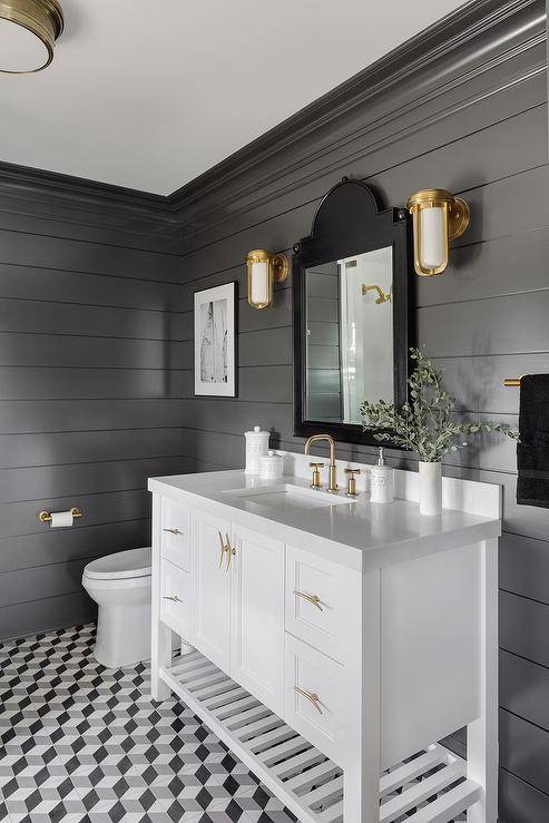 White glass and brass sconces are mounted flanking a black French vanity mirror hung over a white bath vanity fitted with a slatted shelf, brass hardware, and an aged brass faucet kit. The washstand sits on taupe and black geometric floor tiles beside a white porcelain toilet fixed under a black and white art piece.
