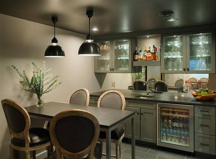 Well appointed gray basement wet bar features gray walls illuminated by two black industrial pendants hung from a dark gray ceiling over an industrial pub table seating four round back French counter stools. Behind the table, lighted glass front cabinets flank a custom lit shelf fixed above a small sink with a polished nickel faucet fixed to a black quartz countertop. The countertop is mounted against a mirrored backsplash and accents gray shaker cabinets finished with oil rubbed bronze hardware and a glass from beverage fridge.