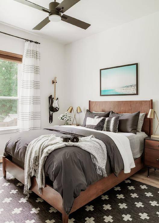 A beach art piece hangs over a brown oak bed complemented with dark gray bedding and gray layered pillows. The bed, placed on a black and white geometric rug, is flanked by two-tone vintage nightstands illuminated by brass task lamps.