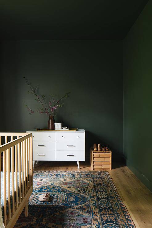 Forest green nursery walls and ceiling complement a white mid-century modern dresser placed behind a pink and blue vintage rug positioned beneath a blond wood crib.