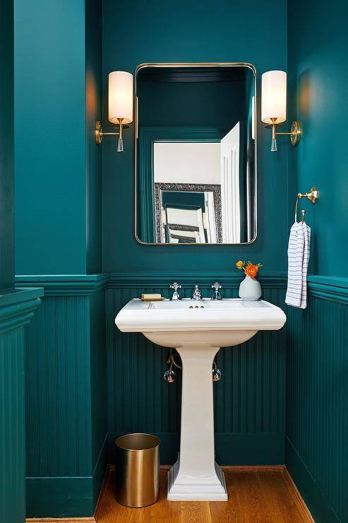 Stylish peacock blue powder room boasts a peacock blue beadboard trim accenting a peacock blue painted upper wall lit by Hudson Valley Lighting Birch Sconces mounted flanking a curved nickel mirror. The mirror is hung above a white porcelain pedestal sink finished with a polished nickel faucet kit.