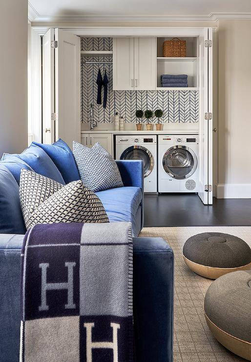 Wonderfully styled family features a blue velvet curved sofa draped in a blue Hermes Avalon blanket and placed on a beige rug facing black and tan poufs. The family room is finished with a washer and dryer fitted in a closet and enclosed beside white cabinets fitted with a sink and polished nickel gooseneck faucet. A clothing rail is fitted over the sink, beneath a white shelf, and beside a white cabinet.