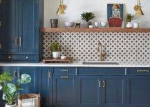 Gorgeous blue kitchen cabinets adorned with brass pulls are finished with an undermount sink and brushed gold gooseneck faucet fixed in front of white and gray mosaic backsplash tiles. The faucet is positioned beneath a brown wood floating shelf lit by two brass swing arm sconces.