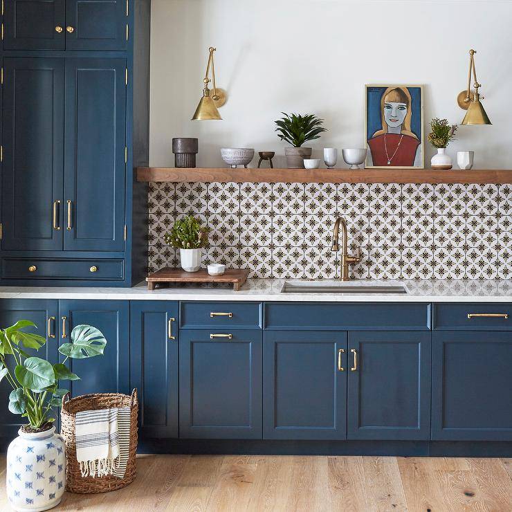 Gorgeous blue kitchen cabinets adorned with brass pulls are finished with an undermount sink and brushed gold gooseneck faucet fixed in front of white and gray mosaic backsplash tiles. The faucet is positioned beneath a brown wood floating shelf lit by two brass swing arm sconces.