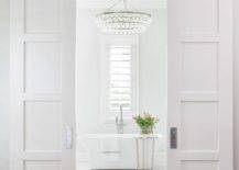 Bi-fold white doors on rails open to a master bathroom lit by a Chandelier hung over an elegant freestanding bathtub placed on white herringbone pattern floor tiles beside accent table and beneath a window.