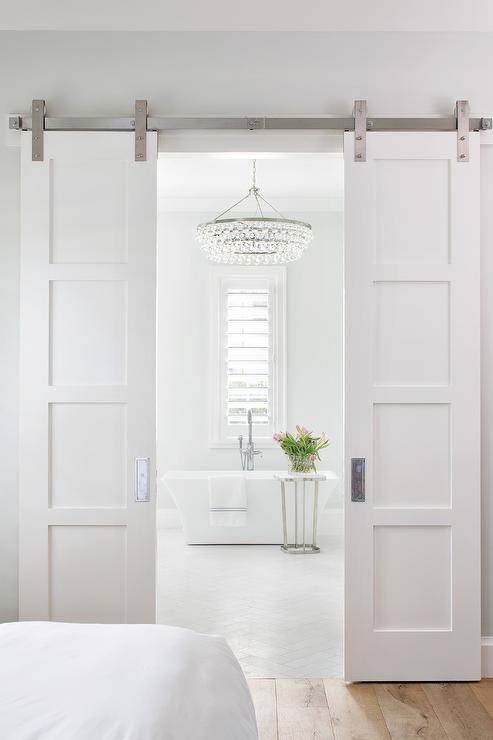 Bi-fold white doors on rails open to a master bathroom lit by a Chandelier hung over an elegant freestanding bathtub placed on white herringbone pattern floor tiles beside accent table and beneath a window.