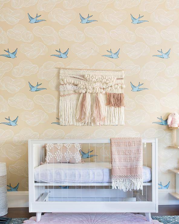 Hygge & West Daydream Wallpaper dresses up a boho baby nursery with a fun and charming finish in beige and blue. Wood and lucite nursery crib features blush pink accents on a throw fringe blanket and an accent pillow.
