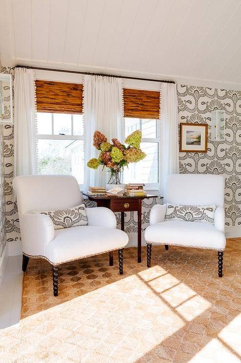 Bamboo roman shades are layered behind white curtains with pom pom trim hung behind white accent chair with black spindle legs placed flanking a dark stained wood accent table positioned on a tan rug. Walls are covered in black and white wallpaper.