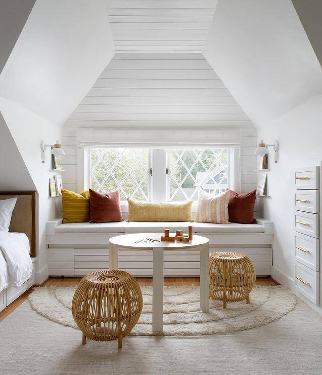 Charming girls' bedroom features rattan stools placed on a cream rainbow rug flanking a round two tone desk. The desk sits in front of a built-in window seat topped with red and yellow pillows and placed beneath a window dressed in a white roman shade and framed by white plank trim.