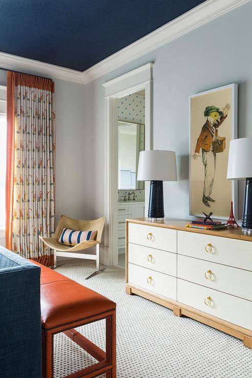 Boy's bedroom features light blue walls and a navy blue painted ceiling, a two-tone white and tan dresser lit by navy blue and white lamps and an orange leather bench at the foot of the bed, atop a tan carpet.