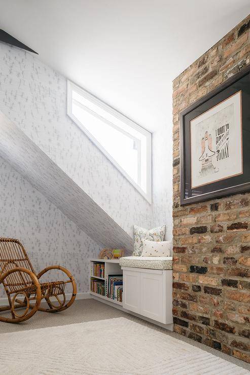 Gender neutral attic nursery features a red brick wall, rattan nursery rocker and a built-in window seat with whimsical pillow.