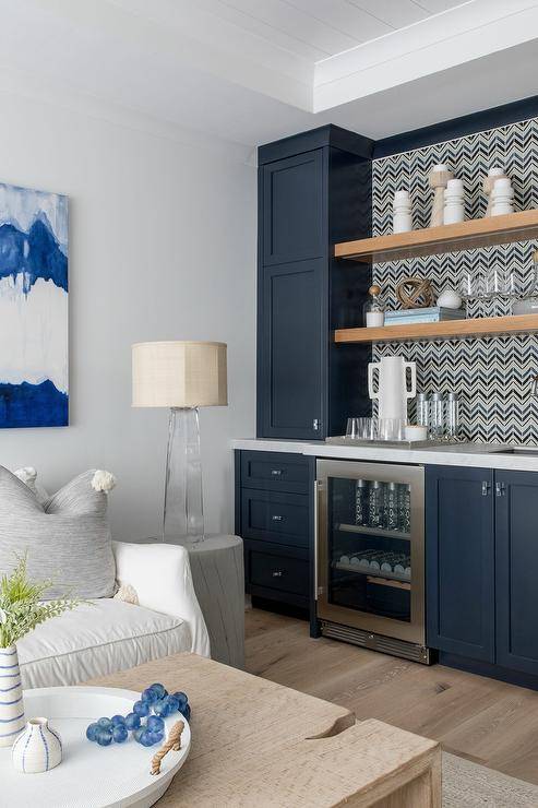 Blue family room wet bar features blue shaker cabinets accented with glass knobs and a marble countertop fixed over a mini glass front beverage fridge. Stacked oak floating shelves are mounted against blue and gray chevron backsplash tiles and between blue upper cabinets.