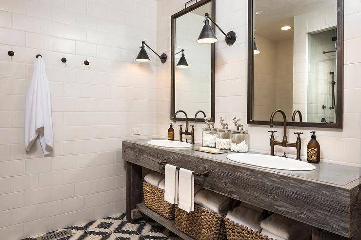 Country style bathroom features wall clad in large white subway tiles fitted with brass towel hooks mounted on a wall adjacent to a reclaimed wood double vanity finished with oval sinks with vintage brass deck mount bridge faucets separated by glass deer canisters and complementing a brass towel bar centered above a shelf holding wicker towel baskets. Above the washstand Boston Functional Library Wall Lights flank the mirrors lighting black and white diamond patterned cement floor tiles.