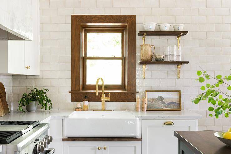 Stained wood moldings frame a window surrounded by white glazed backsplash tiles and positioned beside wooden shelves with brass brackets. The window is also located above a farmhouse sink matched with a brushed gold gooseneck faucet mounted over white cabinets with brass knobs.