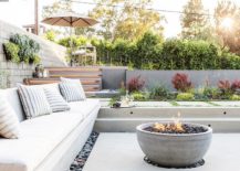 Sunken patio is filled with a concrete bench lined with white outdoor cushions and gray striped outdoor pillows faces a concrete bowl fire pit surrounded by black river rocks.