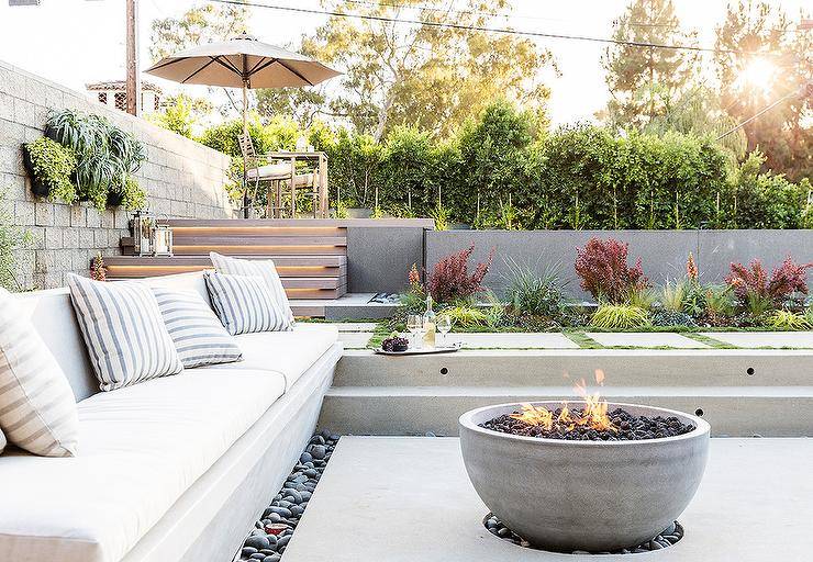 Sunken patio is filled with a concrete bench lined with white outdoor cushions and gray striped outdoor pillows faces a concrete bowl fire pit surrounded by black river rocks.
