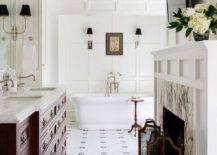 Elegant master bathroom is fitted with a fireplace boasting a gray marble surround framed by a whtie mantel fixed beneath a small TV. The TV is fixed to a wainscot wall facing a cherrywood bath vanity donning vintage hardware and a honed marble countertop
