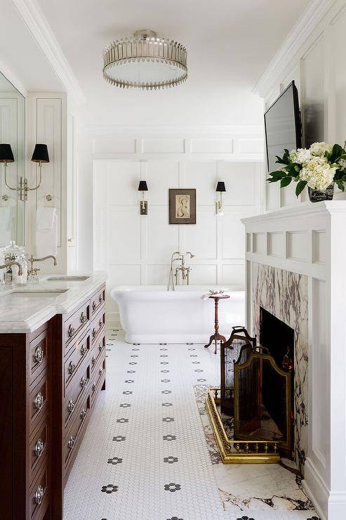 Elegant master bathroom is fitted with a fireplace boasting a gray marble surround framed by a whtie mantel fixed beneath a small TV. The TV is fixed to a wainscot wall facing a cherrywood bath vanity donning vintage hardware and a honed marble countertop