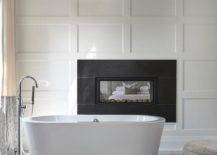 Modern oval tub in front of a black fireplace surrounded by a board and batten wall in a master bathroom design.