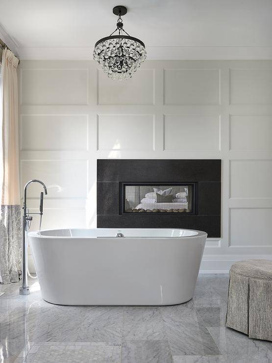 Modern oval tub in front of a black fireplace surrounded by a board and batten wall in a master bathroom design.