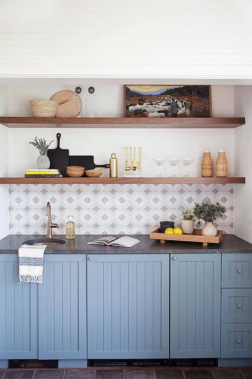 Brown wooden floating shelves are stacked above white and gray mosaic tiles mounted above blue breadboard cabinets donning a black leather countertop. A round hammered brass sink is matched with a satin nickel gooseneck faucet.