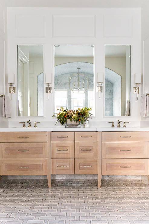 Tall mirror panels lit by are fitted over a blond wood dual washstand placed on gray carrera marble maze tiles. The washstand is adorned with polished nickel pulls and a white quartz countertop fitted with Brizo Rook Widespread Bathroom Faucets with Pop-Up Drain Assembly.