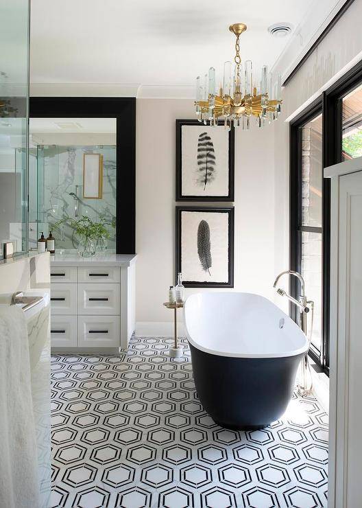A lucite and brass chandelier lights a black and white cast iron bathtub placed on black and white hexagon floor tiles and paired with a floor mount chrome tub filler. The tub filler is mounted in front of black framed windows, while stacked feather art in black frames is hung behind the bathtub.