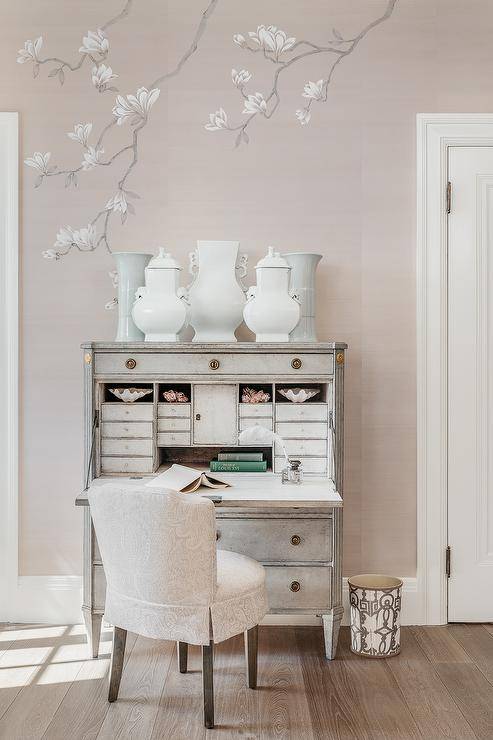 A pink damask chair sits at a gray French drop down desk topped with white and gray vases accenting pink wallpapered walls.