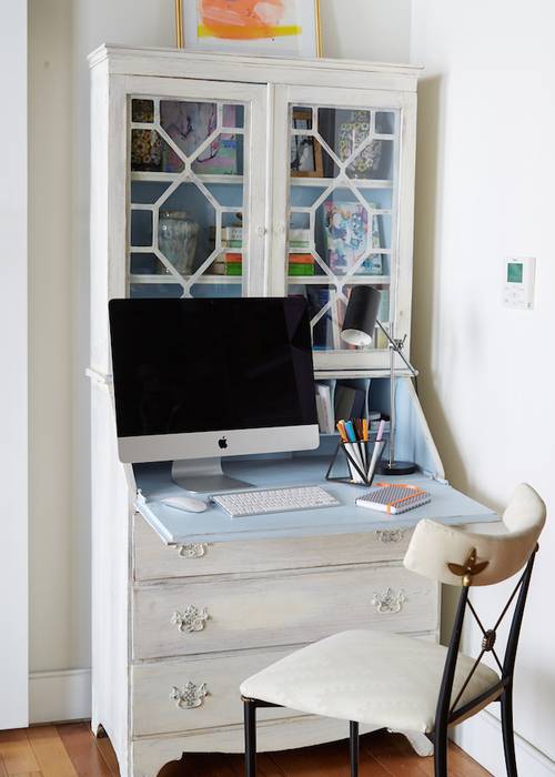 A white and black French desk chair sits in front of a white French secretary desk accented with a blue interior and fitted with a pull down desk top and glass front cabinets.