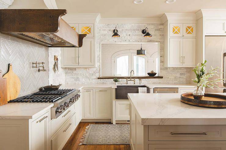 Cottage kitchen design features white stone ledge wall tiles, a French hood over a stainless steel oven, white kitchen cabinets and a white kitchen island topped in gray marble.