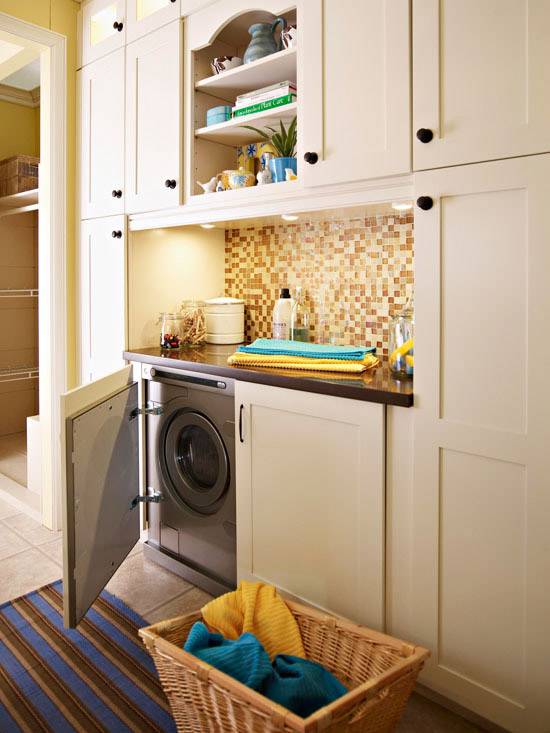 Gorgeous laundry room with silver front-load washer & dryer hidden behind ivory cabinets, mosaic glass tiles backsplash and striped blue & brown rug.