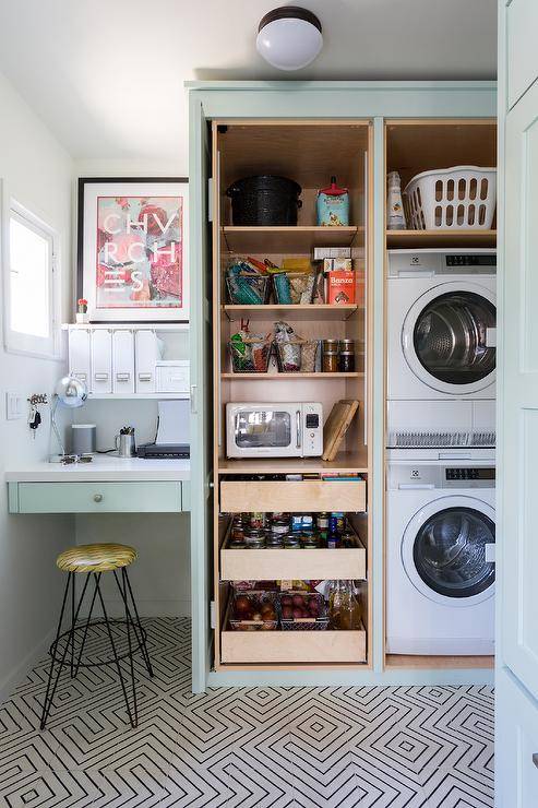 Well-appointed mint-green contemporary laundry room and pantry combo features pantry shelves and pull out drawers hidden beside a stacked washer and dryer and behind mint green cabinet doors. Black and white cement floor tiles lead to a hairpin stool placed at a mint-green built-in desk mounted beneath styled white floating shelves.