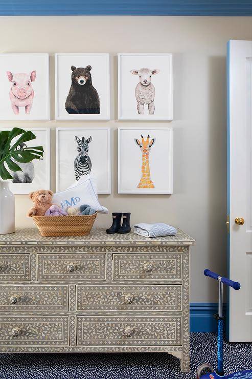 Bright blue crown moldings and baseboards accent an ivory painted wall holding a gallery of framed baby animal sketches over a cream and black bone inlay dresser in a wonderfully styled nursery.