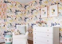 Girl's nursery features pink blue green yellow and gray wallpaper, a white nursery dresser with brass knobs and an ivory roll arm glider over a cream rug.