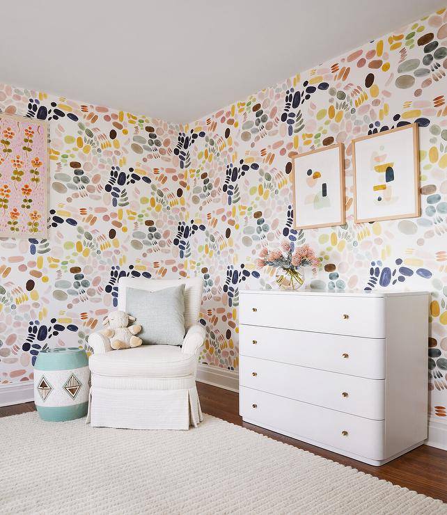 Girl's nursery features pink blue green yellow and gray wallpaper, a white nursery dresser with brass knobs and an ivory roll arm glider over a cream rug.