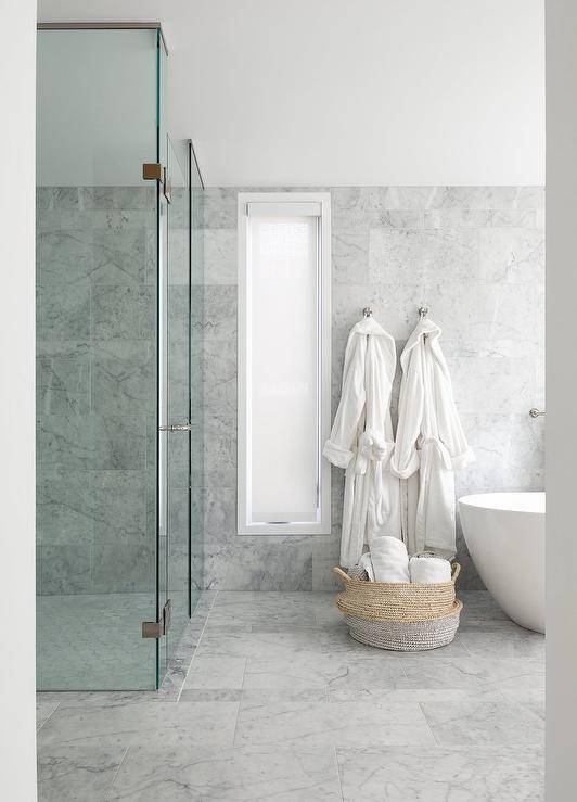 Spacious marble bathroom features his and hers robe hooks over a woven basket on gray marble tiles and a walk in shower with clear partition.