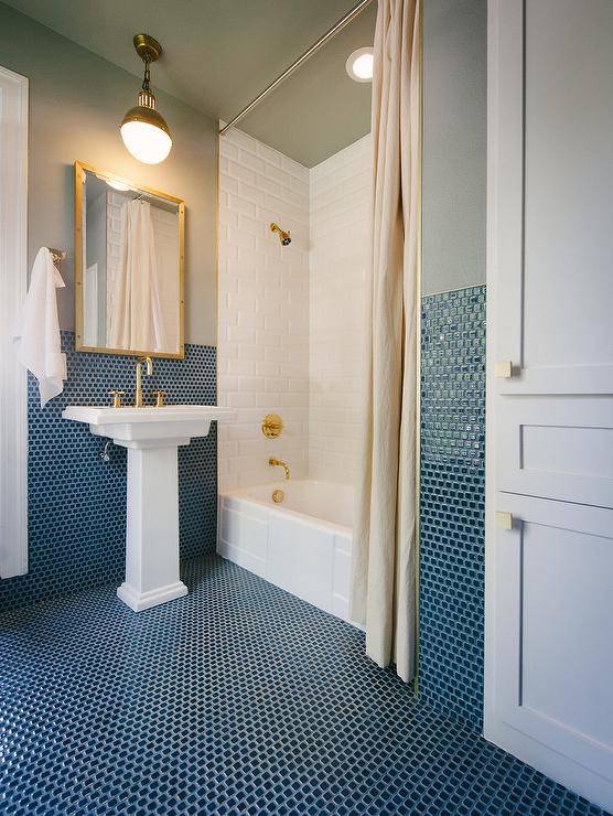 Ocean shade of blue mini grid tiles adds a spa-like tranquility to this bathroom while accented with gold hardware. A Kohler Tresham Pedestal Bathroom Sink with a Purist Brass faucet complements a white drop in bathtub with a polished brass shower kit. Navy blue mosaic tiles make their way up walls providing a seamless appeal illuminated by a brass Hicks Pendant light. Under the pendant, a brass rivet medicine cabinet from Restoration Hardware hangs over matching brass sink accents. This gorgeous reflective bathroom surface, a white storage cabinet closet is a contrast featuring a set of square brass door knobs.