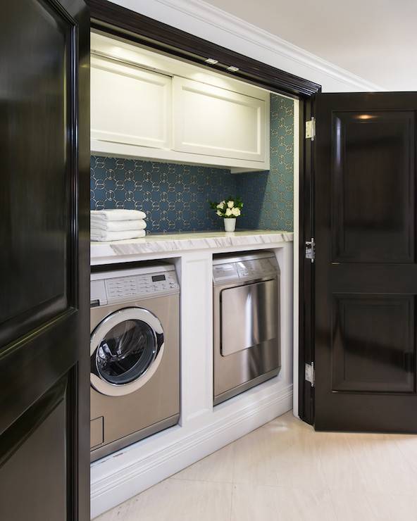 Glossy black double doors open to reveal cabinets suspended over Hermes Fil d'Argent Wallpaper enclosed front load silver washer and dryer topped with white marble.