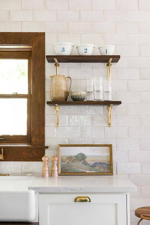 Wood and brass shelves are stacked and mounted against white glazed backsplash tiles beside a window and over white kitchen cabinets donning a brass cup pulls.