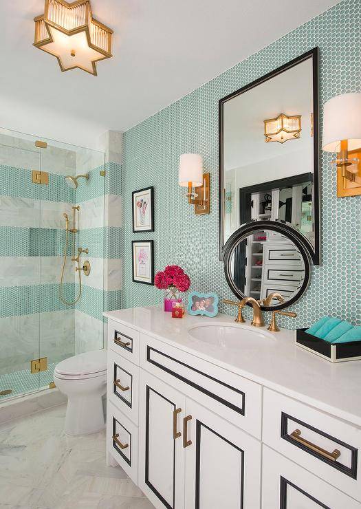 White herringbone marble tiles cover the floors of this exquisite contemporary bathroom featuring a white and black washstand accented with brass pulls and an aged brass faucet topped with a white quartz countertop placed under an upside down black framed mirror illuminated by a Sophia Small Flush Mount and brass sconces mounted to a wall covered in turquoise penny tiles. A seamless glass shower fitted with an aged brass shower kit and lined with stripes of turquoise penny tiles and white marble shower tiles is positioned beside two black framed prints hung over a simple ceramic toilet.