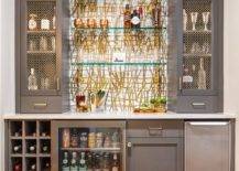 Gold and gray wet bar features floating glass bar shelves, a built in wine rack, charcoal gray cabinets accented with brass pulls and brass grille bar cabinet doors.