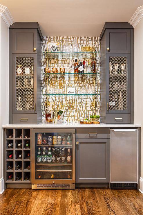 Gold and gray wet bar features floating glass bar shelves, a built in wine rack, charcoal gray cabinets accented with brass pulls and brass grille bar cabinet doors.