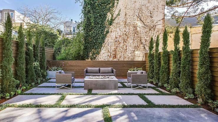 Patio features large concrete pavers with grass trim, a concrete rectangular fire pit, a gray outdoor sofa with gray cushions and a wooden privacy fence.