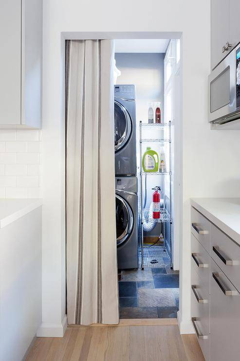 Separated from the kitchen by a gray curtain hung from a doorway, a modern laundry room boasts a stacked silver front loading washer and dryer positioned beside a stainless steel industrial shelving unit.