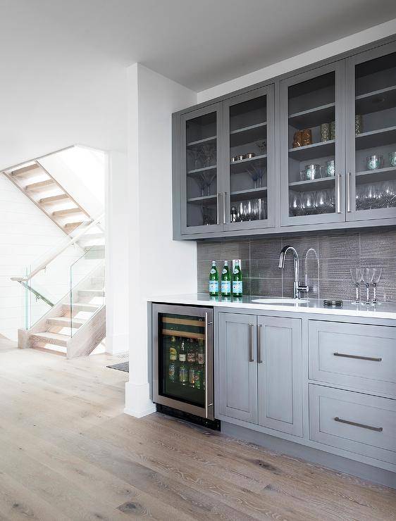 Fixed over gray stacked backsplash tiles, glass front cabinets are located over gray shaker cabinets donning polished nickel hardware and fitted with a glass front beverage fridge and a sink with a polished nickel gooseneck faucet.